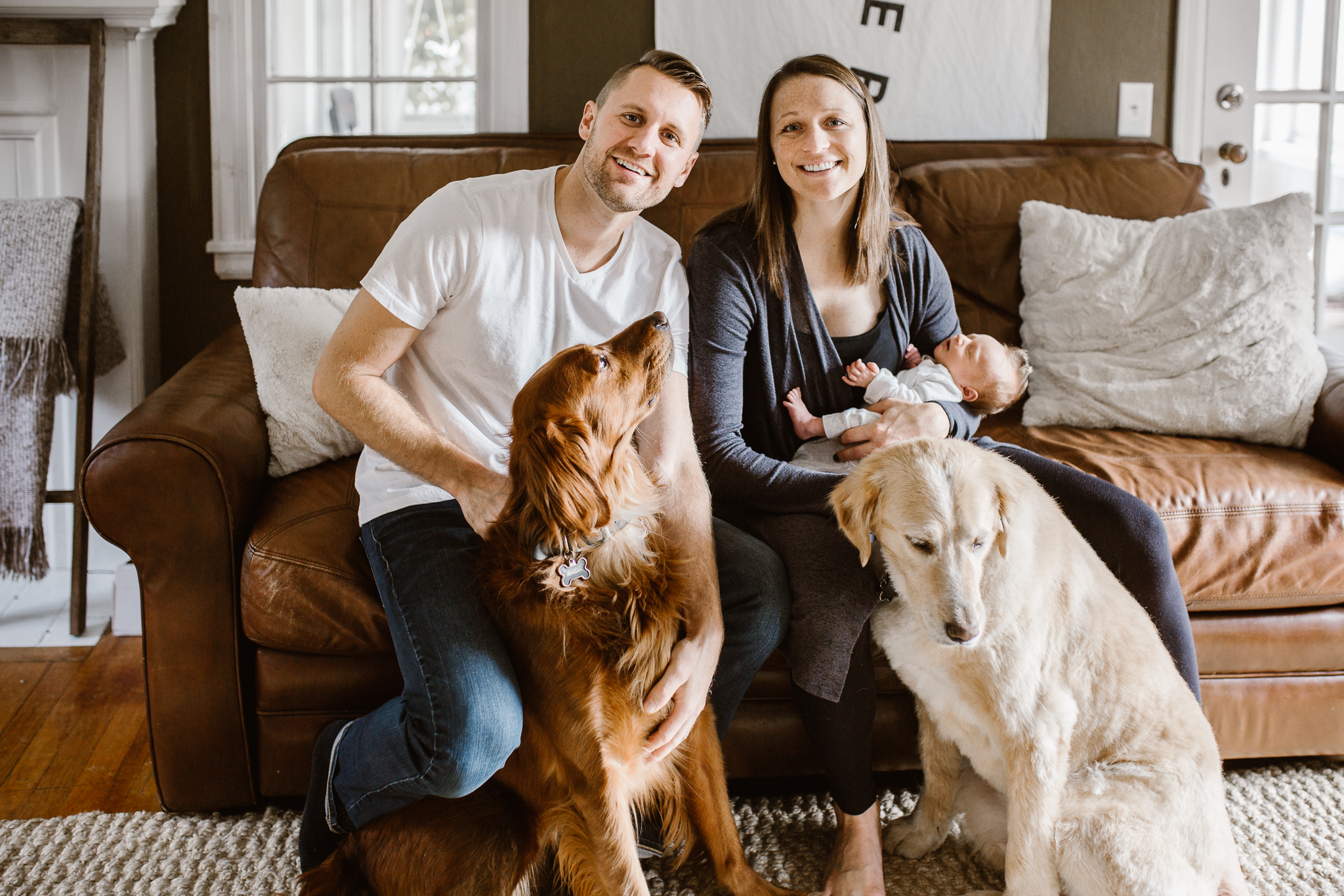 New parents holding thier baby on the couch along with their two dogs.
